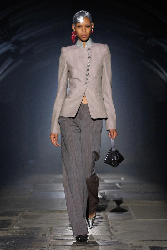 Modelka w eleganckim, szaro-prążkowanym garniturze, trzymająca stylową torebkę, idzie po wybiegu w dymnym oświetleniu.