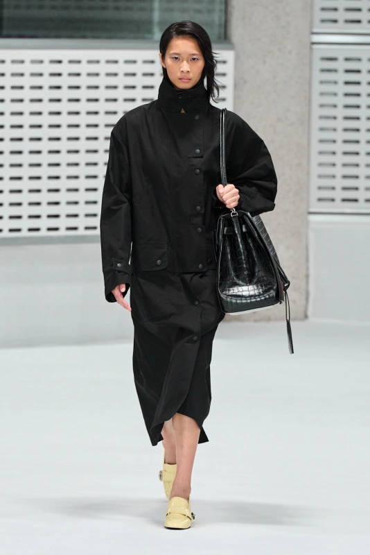 Model w czarnym, oversize'owym płaszczu trzyma czarną torebkę w stylu tote.