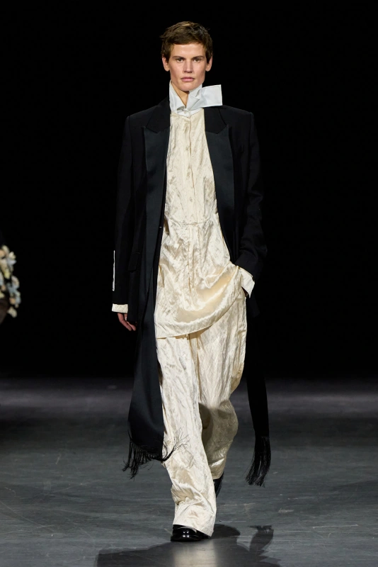 Model w eleganckim, ciemnym płaszczu i jasnej, jedwabnej odzieży, prezentujący modę na wybiegu.