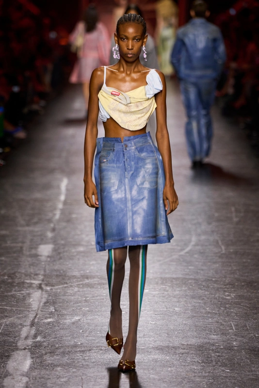 Modelka prezentuje niebieską spódnicę z denimu oraz materiałowy top, na tle wybiegu mody.