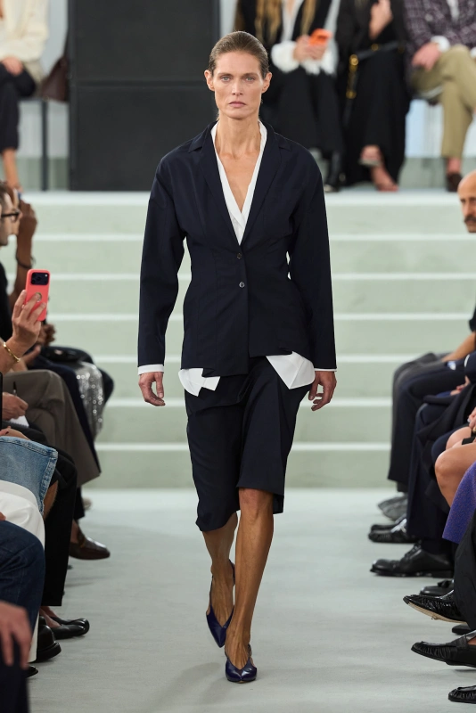 Modelka na wybiegu ubrana w elegancki, ciemny garnitur z białą koszulą w stylu oversize. W tle widownia.
