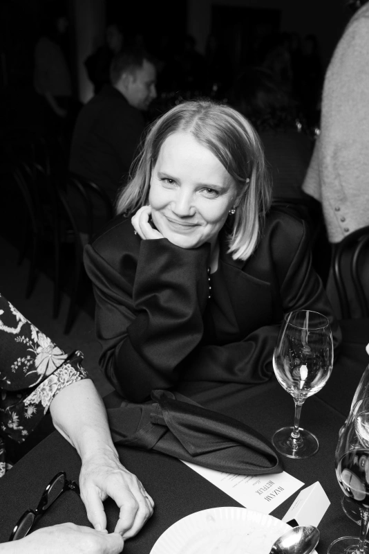 Joanna Kulig z krótkimi włosami przy stoliku, uśmiechająca się i opierająca podbródek na dłoni. W tle widać innych gości.
