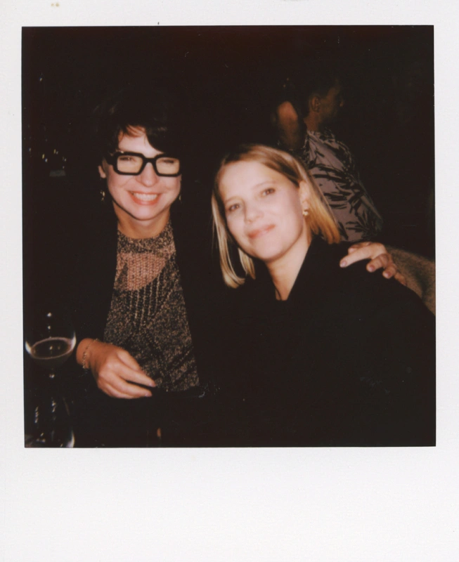 Joanna Kulig siedzi obok stylowo ubranej kobiety, uśmiechając się do kamery w eleganckim otoczeniu.