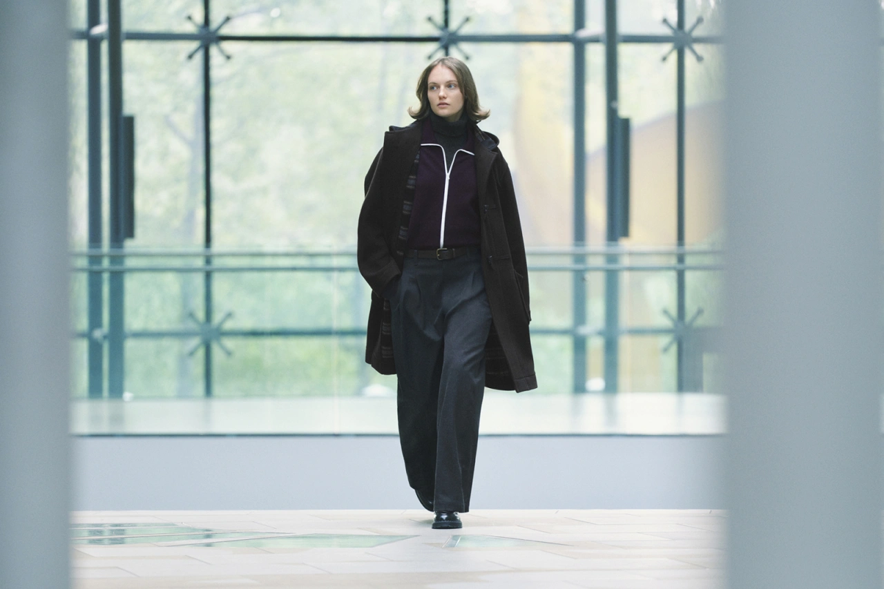 Modelka w eleganckim, ciemnym płaszczu oraz szerokich spodniach, przechadza się w nowoczesnej przestrzeni.