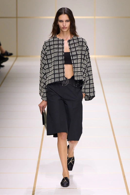 Modelka w eleganckim zestawie: krótki żakiet w kratę, czarny top i spódnice do kolan. Trzyma torebkę.