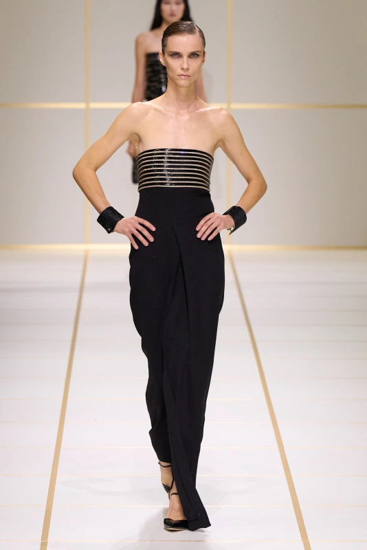 Modelka prezentuje elegancką, czarną suknię z paseczkami w kolorze złota, podkreślającą sylwetkę. Stylizację uzupełniają czarne bransoletki.