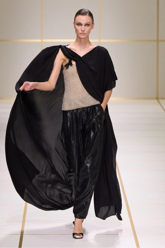Modelka w eleganckiej stylizacji, z błyszczącą bluzką i czarnymi, luźnymi spodniami, prezentująca się na wybiegu.