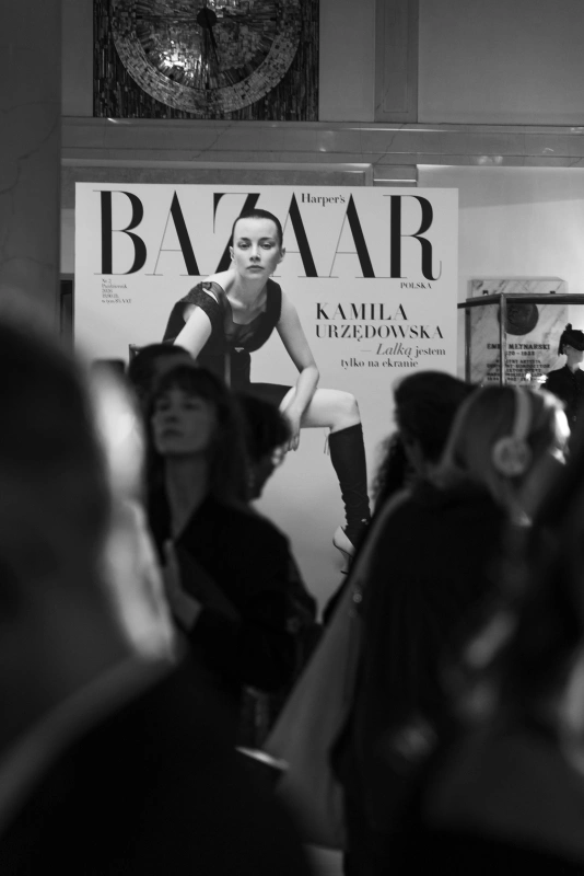 Na pierwszym planie widać postacie ludzi, a w tle znajduje się okładka Harper's Bazaar Polska z Kamila Urzędowską.