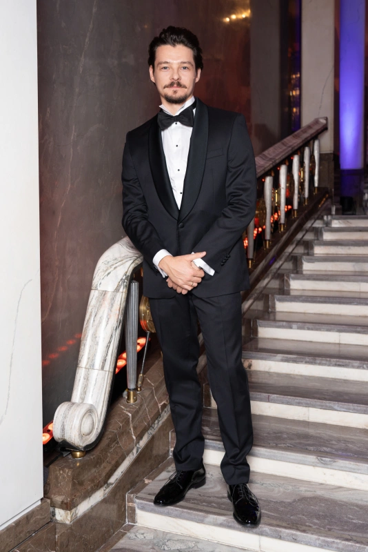 Tomasz Ziętek w eleganckim czarnym garniturze stoi na schodach, przy balustradzie w stylowym otoczeniu.