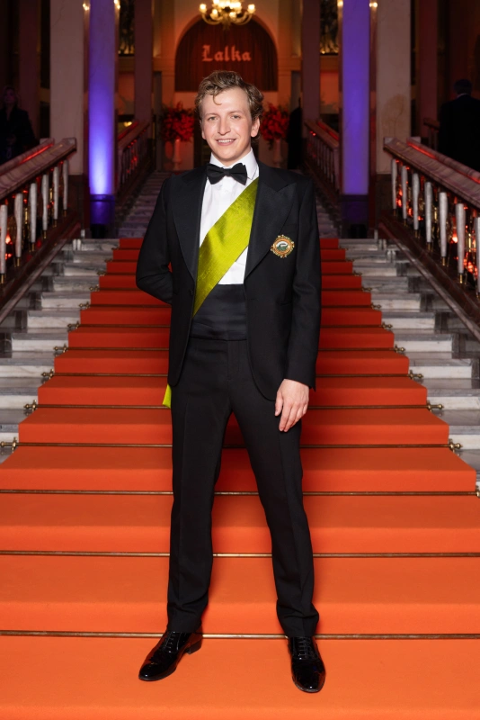 Maciej Musiałowski stoi na tle czerwonych schodów, ubrany w elegancki strój wizytowy z zieloną szarfą.