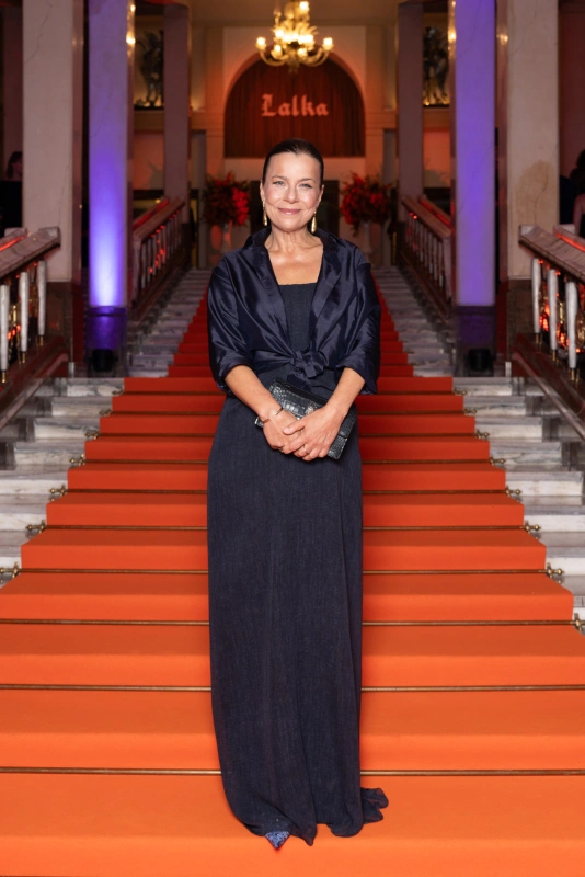 Agata Kulesza w eleganckiej sukience, stoi na schodach z czerwonym dywanem, trzymając kopertówkę.