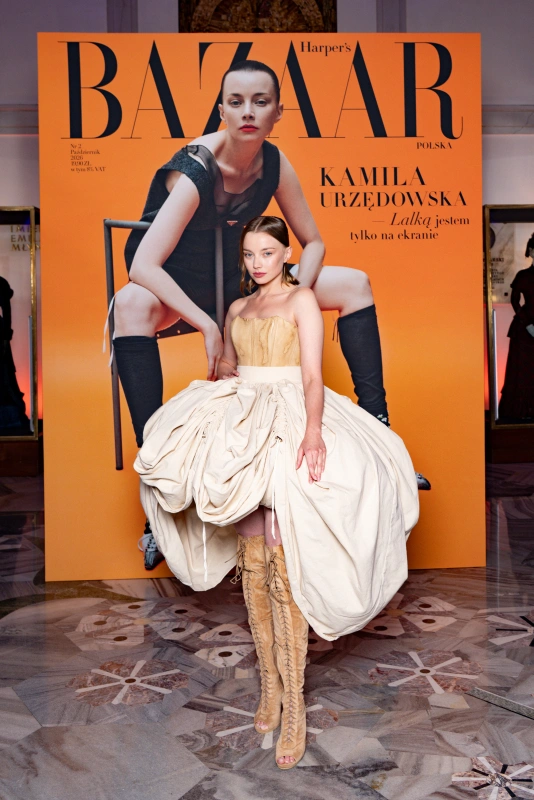 Kamila Urzędowska pozująca na tle okładki "Harper's Bazaar", w eleganckiej sukni i wysokich botkach.