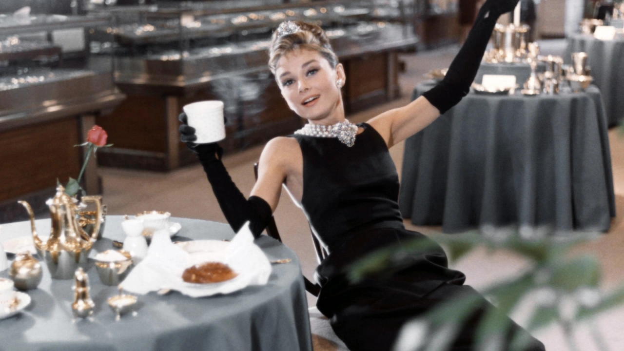 Kobieta w eleganckiej czarnej sukni siedzi przy stole w restauracji, trzymając filiżankę. W tle stoi wystawa srebrnych naczyń.
