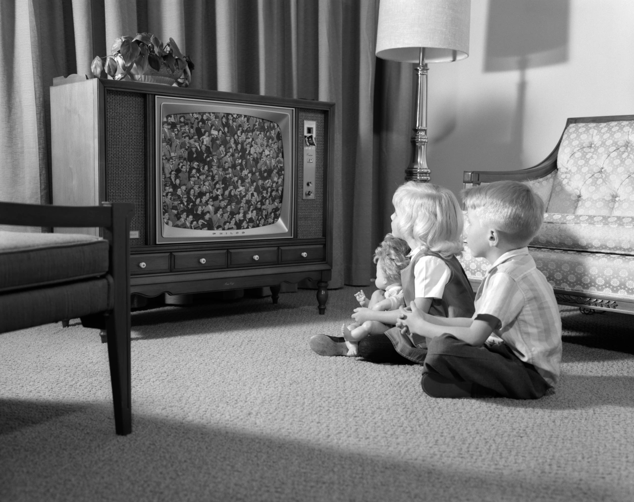 Dwoje dzieci siedzi na podłodze, oglądając telewizję, w otoczeniu mebli w stylu retro.