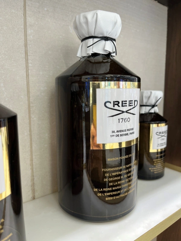 Perfumy Creed w eleganckiej butelce, z białą pokrywką i złotymi detalami, eksponowane na półce.