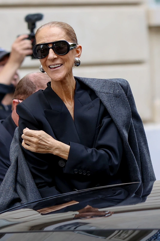 Celine Dion w Paryżu, uśmiechnięta, w eleganckim czarnym stroju i dużych okularach przeciwsłonecznych.