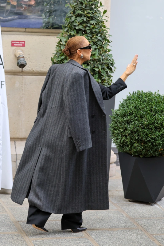 Celine Dion w Paryżu, ubrana w długi, szary płaszcz, wita kogoś na ulicy.