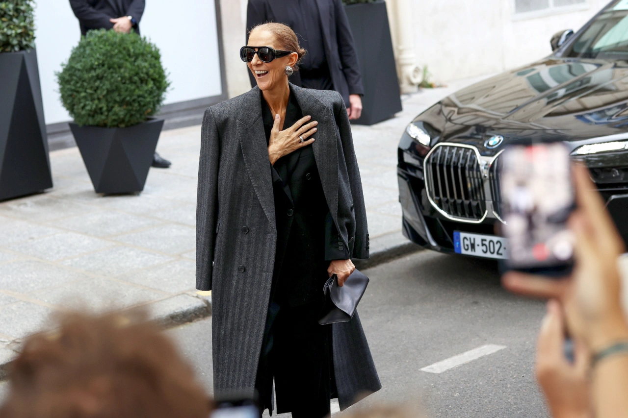 Celine Dion w Paryżu, uśmiechająca się w eleganckim płaszczu, stoi obok luksusowego samochodu i ludzi robiących zdjęcia.