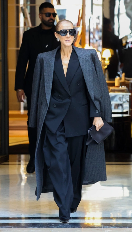 Celine Dion w Paryżu, stylowa w ciemnym garniturze i płaszczu, przechodzi przez eleganckie wnętrze.