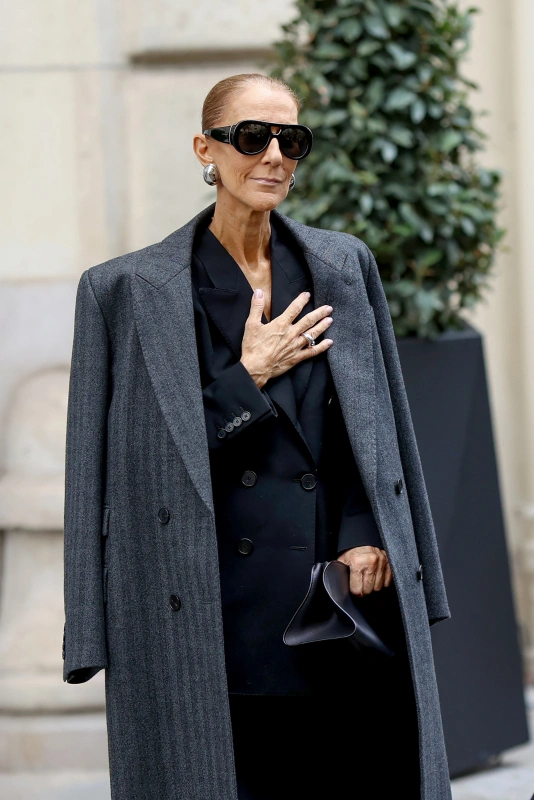 Celine Dion w Paryżu, ubrana w elegancki, szary płaszcz, okulary przeciwsłoneczne i stojąca w przyjaznej pozie.