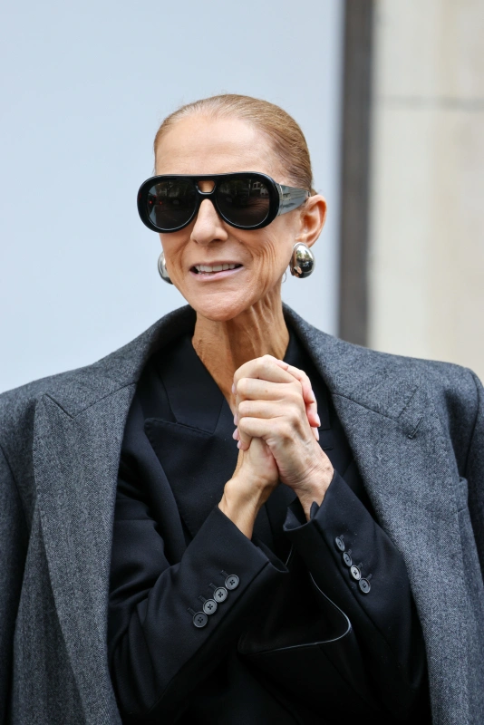 Celine Dion w Paryżu, uśmiechnięta, w eleganckim stroju i dużych okularach przeciwsłonecznych.