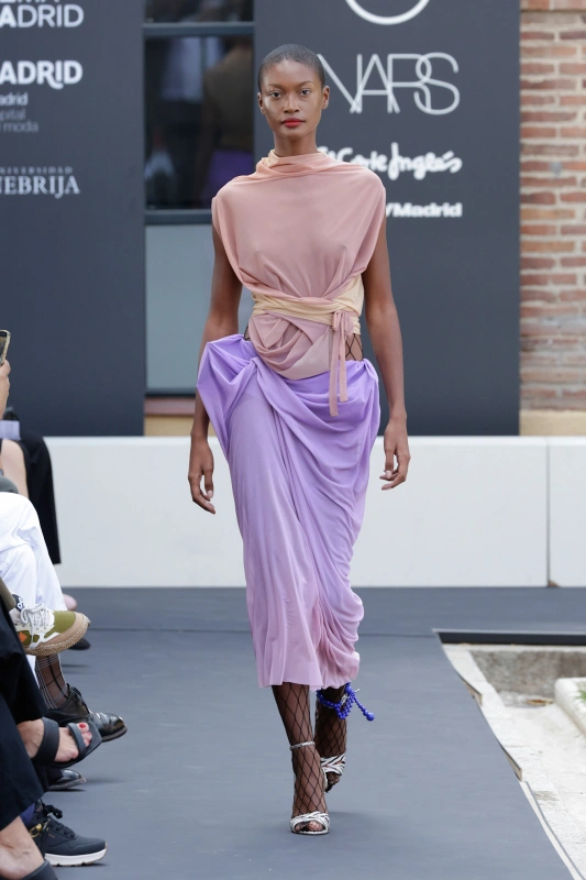 Modelka na wybiegu w eleganckiej, różowo-fioletowej sukni z paskiem. Świetna atmosfera, widownia obserwuje pokaz mody.