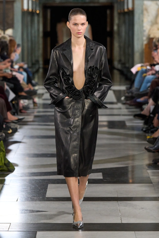 Modelka prezentuje elegancki, czarny płaszcz z kolekcji Simone Rocha wiosna-lato 2027, z efektownymi detalami.