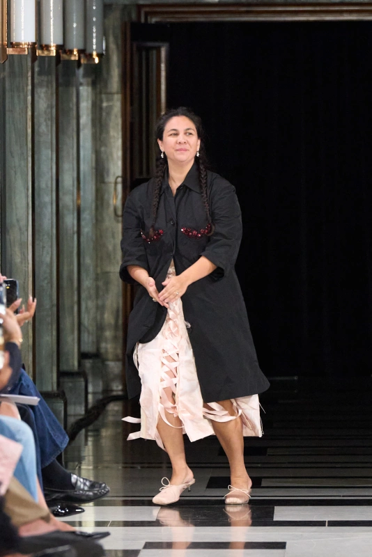 Simone Rocha prezentuje swoją kolekcję na wybiegu, ubrana w czarny płaszcz i jasną sukienkę.