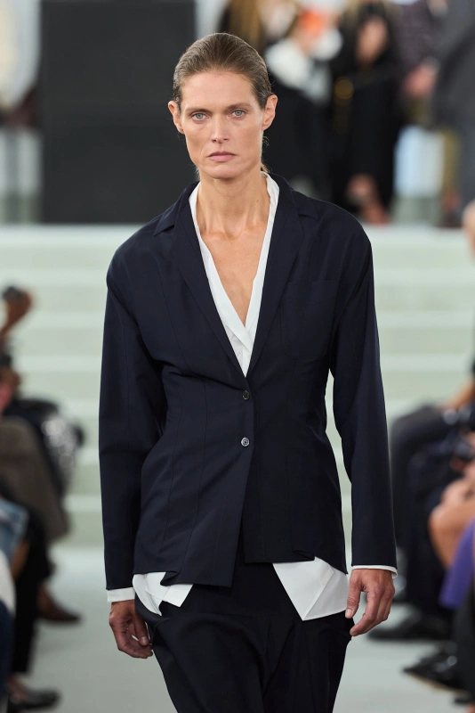 Pokaz Jil Sander prezentuje modelkę w eleganckim, ciemnym kostiumie na tle publiczności.