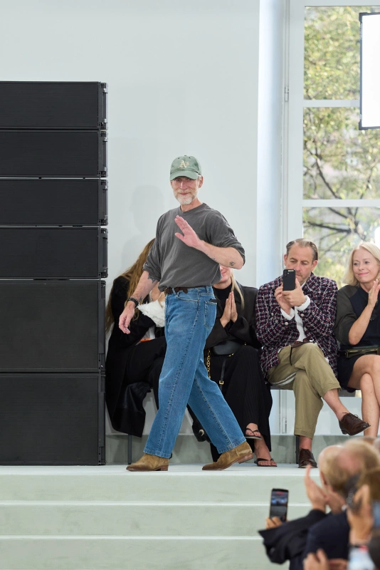 Pokaz Jil Sander, na którym widoczna jest osoba w szarej koszulce i niebieskich dżinsach, machająca do widowni.
