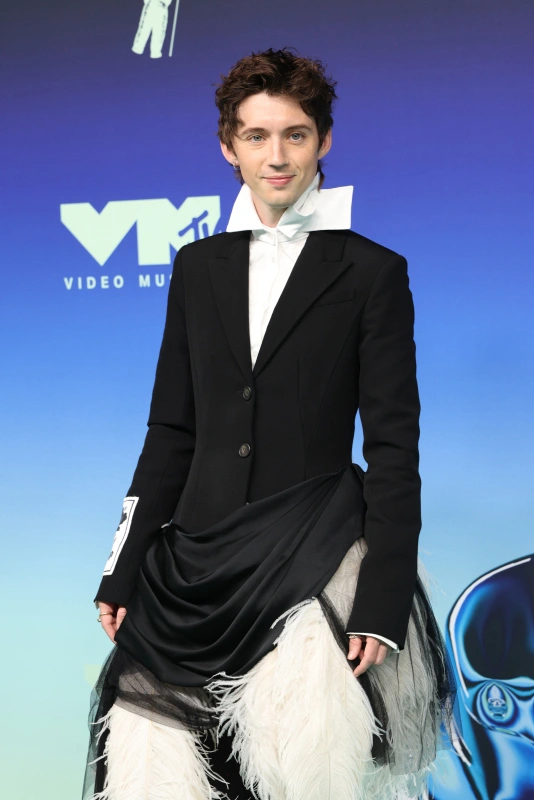Troye Sivan w eleganckim, nietypowym stroju, z białym kołnierzykiem i elementami ozdobnymi. Tło w kolorach niebieskich.